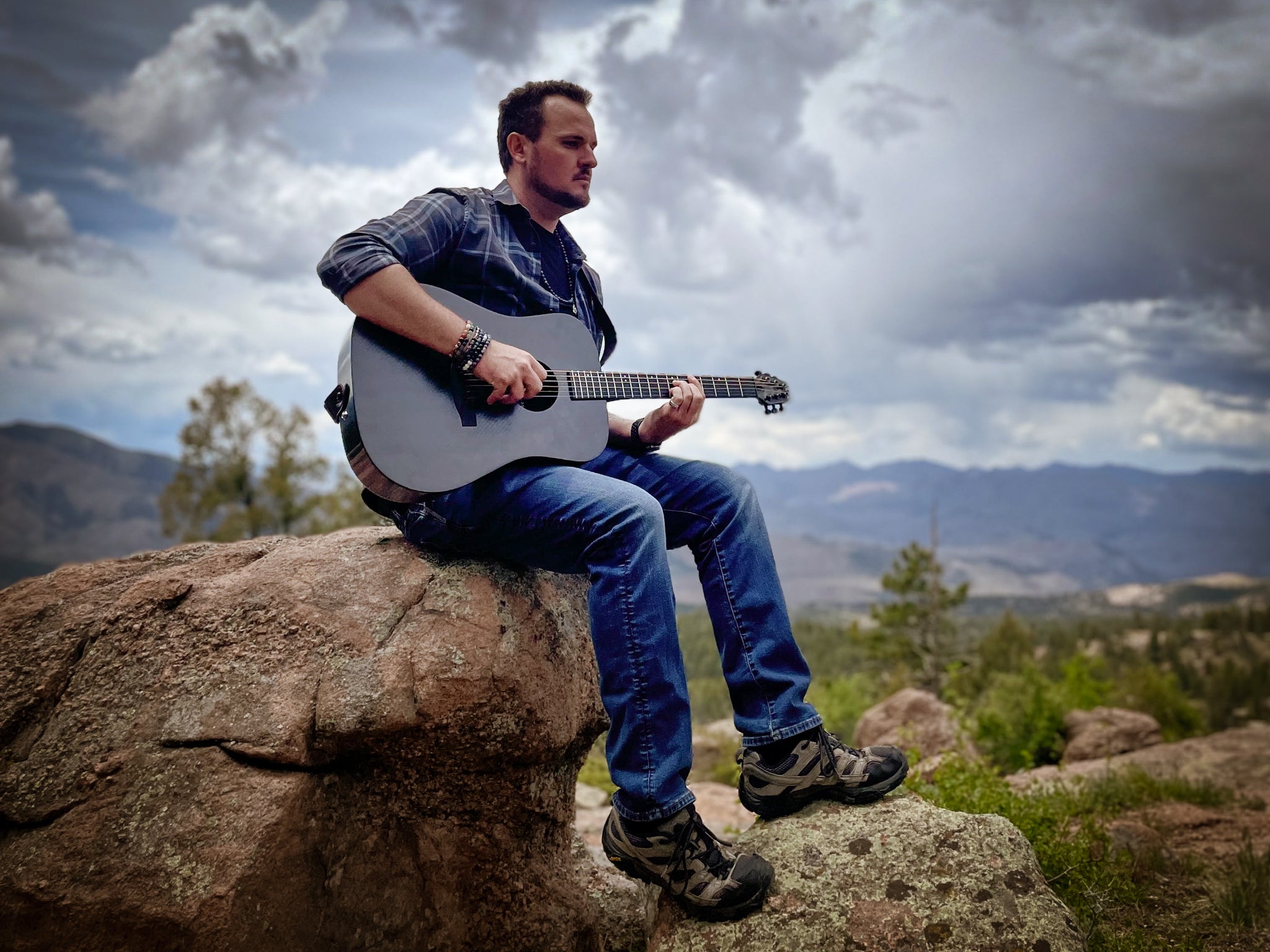 A man rests on a boulder while playing the KLŌS Full Carbon Full Size Guitar 