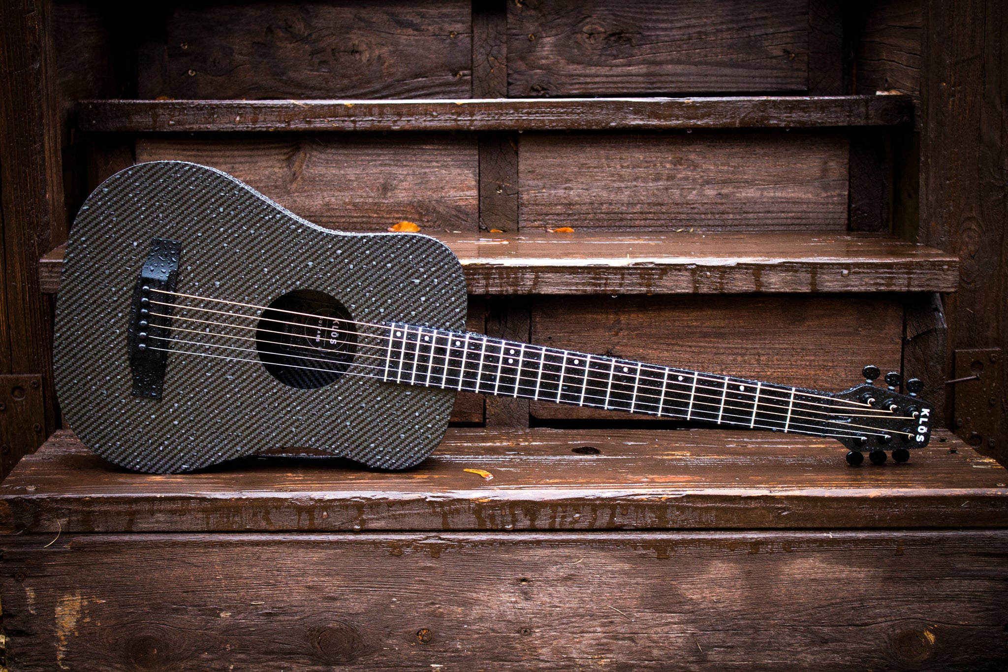 A Full Carbon Travel guitar rests on wooden steps while rain pours down 