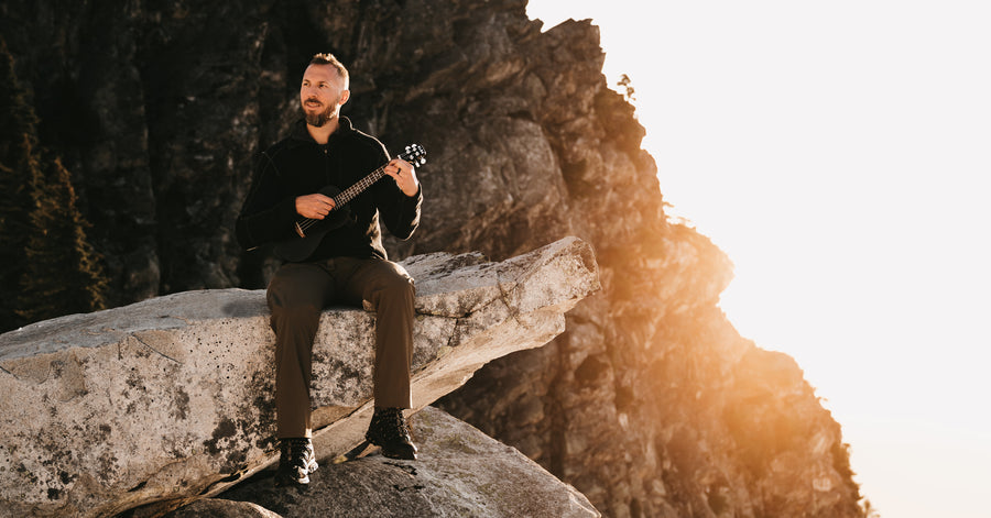 A man is playing a KLŌS tenor uke while sitting on a cliffside 