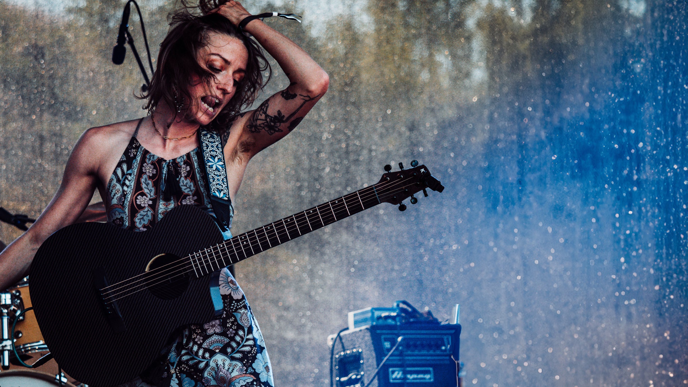 A female musician with tattoos and a face tattoo, wearing a patterned dress, passionately playing an acoustic guitar on stage with a blue-hued spotlight and a visible amp in the background.